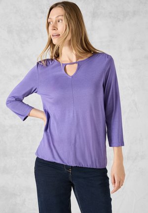 Lila långärmad topp med keyhole-neckdetalj, mjukt tyg och rynkad fåll. Matchas med mörka denimbyxor.