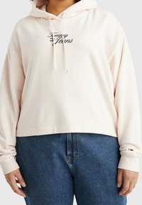 Hoodie cropped di colore rosa chiaro realizzato in tessuto morbido, con logo ricamato nero e cordini. Indossato con jeans in denim blu.