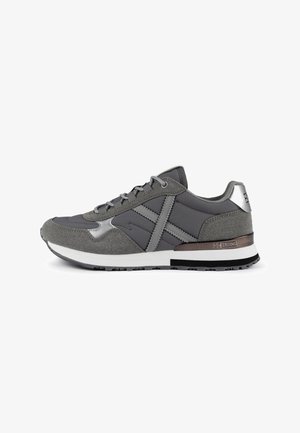 Chaussure de sport grise présentant un mélange de matériaux lisses et texturés, avec des accents métalliques et une semelle blanche rembourrée.