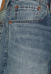 Ljust blå denimjeans med kopparnitar, orange sömmar och en fem-fickorsdesign. Slät textur; synliga bleknings- och whiskeringdetaljer.