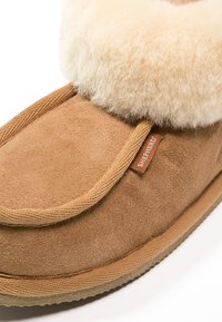 Pantofola in suede color tan con un morbido risvolto color crema, caratterizzata da una trama intrecciata, suola marrone e una piccola etichetta marrone sul lato.