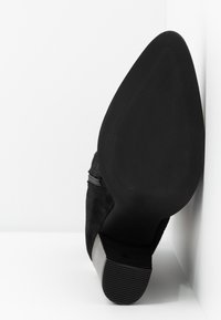 Bottine en daim noir à bout pointu avec un talon bloc, semelle face avant contre un mur et un sol blancs.