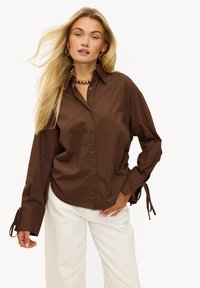 Chemise marron à boutons avec une coupe décontractée, dotée de manches longues avec des liens aux poignets. Portée avec un pantalon blanc à jambes larges.