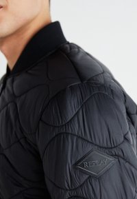 Giacca bomber nera trapuntata con colletto a coste. Presenta motivi cuciti e un'etichetta in gomma sulla manica. Texture liscia.