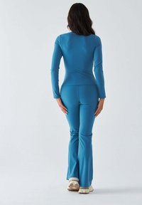 Dopasowany dres z długim rękawem w jednolitym kolorze morskim, wykonany z elastycznego, gładkiego materiału. Widoczne są rozszerzane nogawki i ozdobne przeszycia.