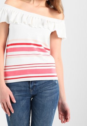 Woman wearing off-shoulder white top with pink and red horizontal stripes and blue jeans against a plain background.