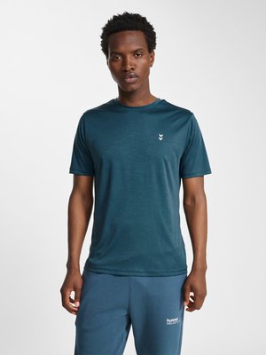 Mann steht und trägt ein türkisfarbenes kurzärmeliges Sportshirt und blaue Hummel Jogginghose vor einem schlichten hellen Hintergrund.