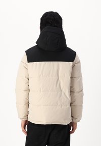 Daunenjacke mit beigem Body und schwarzer Kapuze, ausgestattet mit horizontalen Steppnähten, elastischen Bündchen und einem lockeren Schnitt.