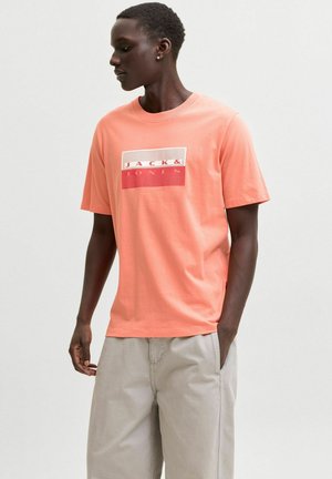 Man wearing coral Jack & Jones t-shirt and light beige pants, standing with one hand in pocket, looking to the side against a plain background.