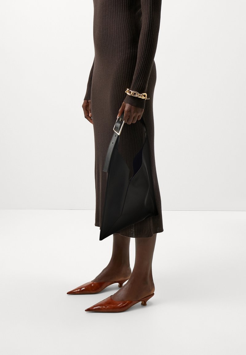 Personne portant une robe marron côtelée, des chaussures marron pointues à petit talon, un bracelet chaîne doré et tenant un grand sac à main en cuir noir.