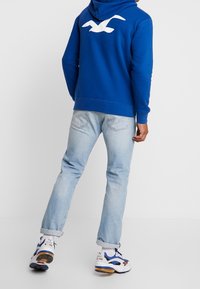 Blue hoodie with a white bird logo on the back, paired with light blue jeans and white sneakers with blue accents.