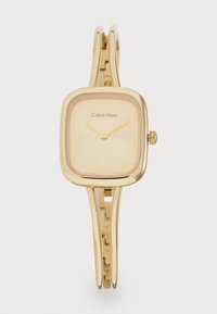 Gold-tone square watch with a smooth dial, slender metal band, and a subtle branding detail. Features a simple crown for adjustment.