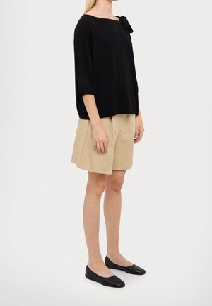 Kvinde stående iført en løs sort top med sløjfedetalje, beige nederdel til knæet og sorte flade sko på en ensfarvet baggrund.