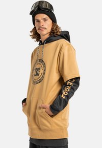 Beżowa, oversize'owa bluza z kapturem z czarnymi akcentami, z dużym logo z przodu i grafiką na rękawie. Posiada kieszeń z przodu i sznurki ściągające.