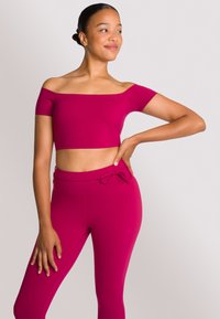 Fuksjowy crop top z dekoltem off-shoulder w połączeniu z legginsami o wysokim stanie z troczkiem. Obie części mają gładką, elastyczną teksturę.