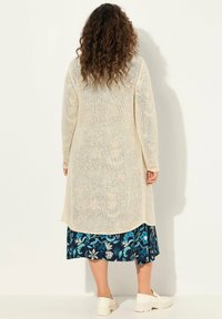 Cardigan lungo e lavorato a maglia aperto di colore crema, caratterizzato da una texture morbida. Indossato sopra una gonna a fiori con dettagli blu e scarpe slip-on bianche.