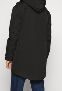 Mand iført en sort hættejakke og blå jeans, vist fra bagsiden mod en ensfarvet lys baggrund.