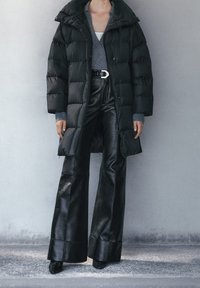 Cappotto puffer nero con colletto alto e chiusura a zip, abbinato a pantaloni in pelle nera a vita alta svasati e maglieria grigia sotto.