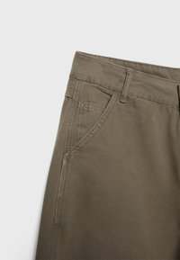 Gros plan d’un pantalon de couleur kaki montrant la ceinture, les passants de ceinture et la poche avant avec des coutures visibles sur un tissu texturé.