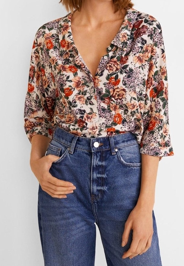 Mujer con blusa de botones de flores y mangas 3/4, metida en unos vaqueros azules de cintura alta, una mano en el bolsillo y la otra relajada.