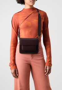 Brun lærkorssbodyveske med to frontlommer, justerbar stropp og lagdelt design. Modellen har på seg en oransje langermet topp og rosa bukser.