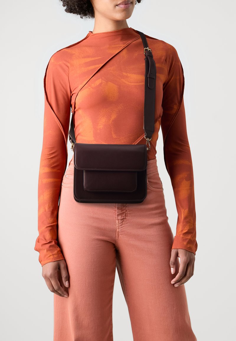 Brun lærkorssbodyveske med to frontlommer, justerbar stropp og lagdelt design. Modellen har på seg en oransje langermet topp og rosa bukser.