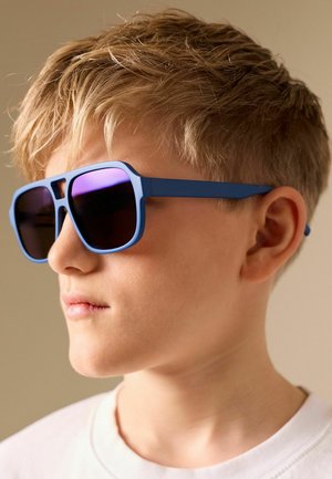 Blondes Kind mit blau gerahmten Sonnenbrillen mit dunklen Gläsern schaut zur Seite vor neutralem Hintergrund.