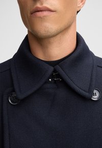 Marineblauwe wollen jas met een hoge kraag, voorzien van een dubbele rij knopen aan de voorkant, knoopaccenten en een gestructureerde stofafwerking.