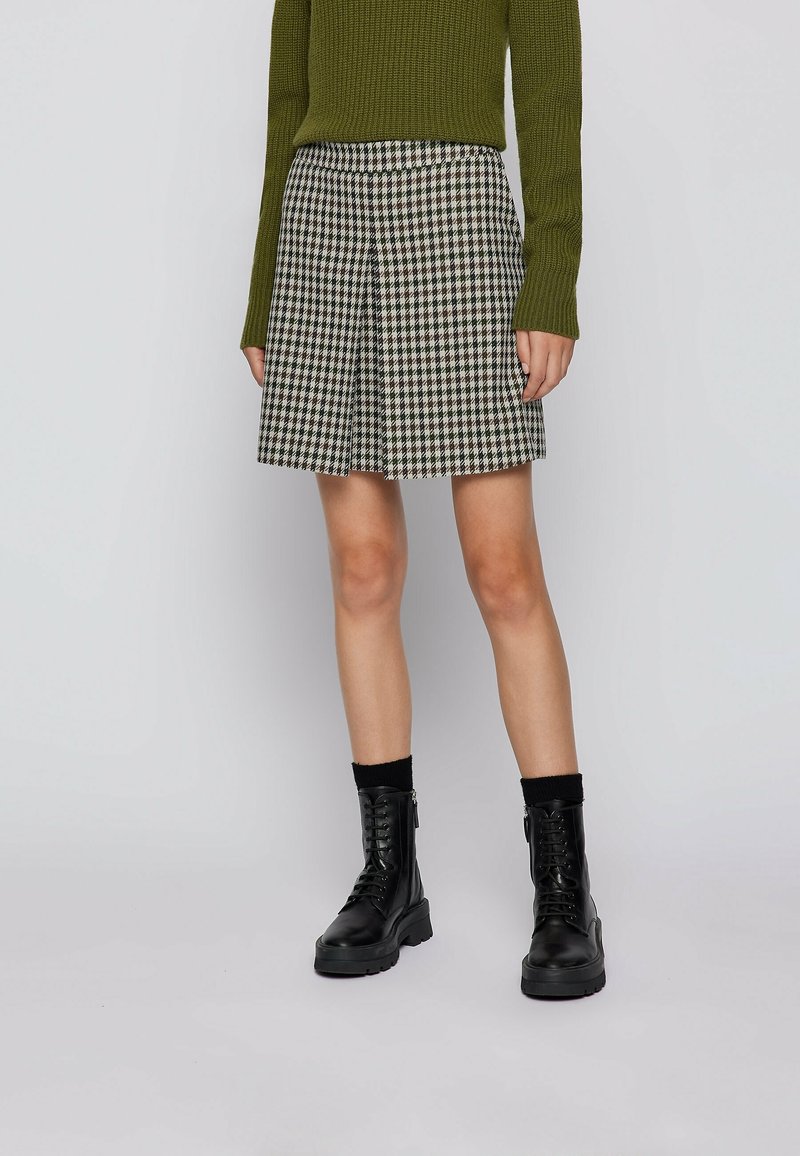 Femme portant une jupe en pied-de-poule noire et blanche, un pull en maille vert olive, des chaussettes noires et des bottines noires à lacets.
