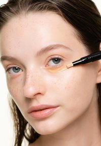 Applicatore di correttore con punta a pennello in uso, che applica un prodotto arancione chiaro sotto l'occhio su una pelle chiara con lentiggini e trucco naturale.