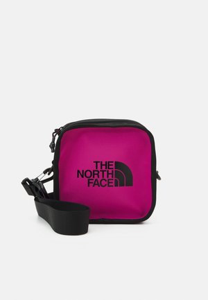 Lille firkantet lyserød crossbody taske med sorte kanter, sort justerbar rem og sort "The North Face" logo foran.