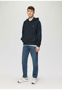 Felpa blu scuro con tasca frontale e dettagli con bottoni, indossata sopra una camicia bianca. Abbinata a jeans blu e sneakers dai toni neutri.