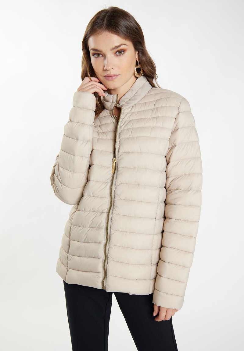 Giubbotto imbottito beige con cuciture orizzontali, colletto alto e dettagli della cerniera dorati. Presenta una texture liscia e isolante. Indossato sopra pantaloni neri.