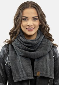 Écharpe en tricot gris foncé avec un motif en côtes texturé, drapée autour du cou, dotée d'une petite étiquette en cuir. Portée avec une veste en cuir noire.
