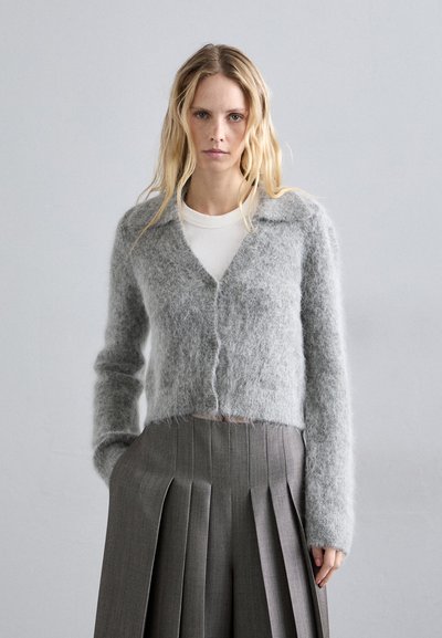 Szürke, bolyhos kardigán V nyakkivágással, gombos záródással és hosszú ujjakkal, fehér felsőre rétegezve, pleated szürke szoknyával párosítva.