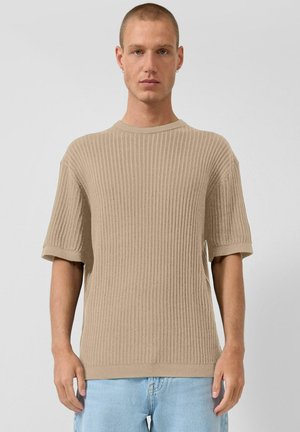 Młody mężczyzna z krótkimi włosami, ubrany w beżowy prążkowany sweter z krótkim rękawem i jasnoniebieskie dżinsy, stoi przodem na jednolitym szarym tle.