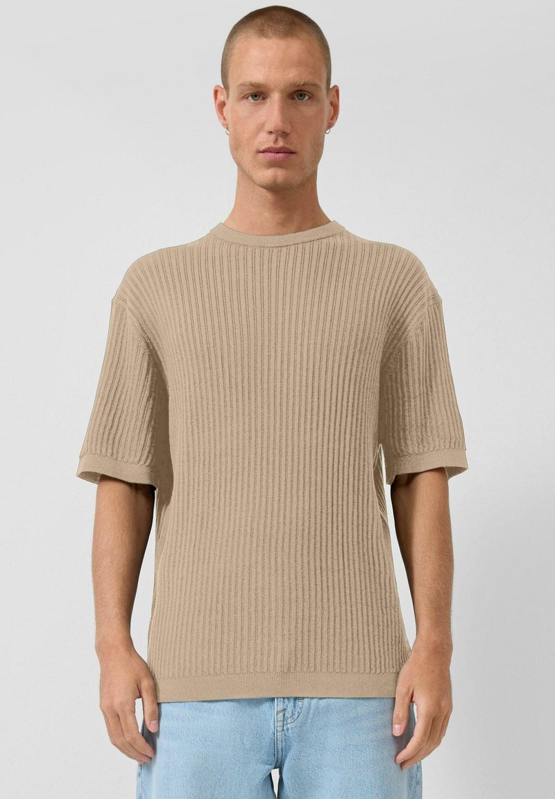 Jeune homme aux cheveux courts portant un pull beige côtelé à manches courtes et un jean bleu clair, face à la caméra sur un fond gris uni.