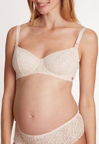 Tehotenská čipkovaná podprsenka v jemnej béžovej farbe s kvetinovým listovým vzorom, nastaviteľnými ramienkami a zapínaním na háčiky. Má jemnú textúru a podporný dizajn.