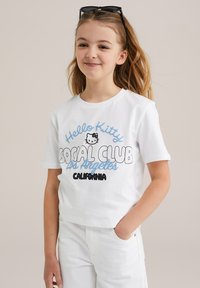 Camiseta de algodón blanca con gráfico de "Hello Kitty" en texto negro y azul, diseño recortado, mangas cortas y escote redondo.