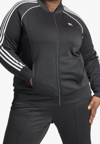 Černá atletická bunda na zip s bílými pruhy na ramenou, vybavená bočními kapsami a hladkou, texturovanou látkou.