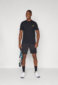 Black athletic t-shirt with short sleeves and a yellow logo, paired with grey shorts. Model holds a tennis racket and wears white sneakers.