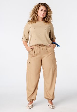 Vrouw met krullend haar die een beige trui met versieringen draagt, losse beige cargobroek, witte gevlochten schoenen en een blauwe sjaalarmband.