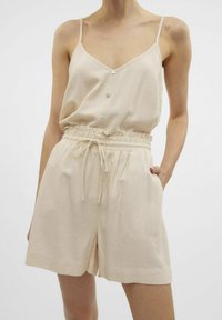 Beige linneoverall med tunna spaghettiband, V-ringning, dekorativa knappar, elastisk midja och sidofickor. Mjukt, texturerat tyg.