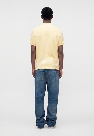Homme debout de dos portant un polo jaune pâle à manches courtes et un jean bleu ample avec des baskets blanches.