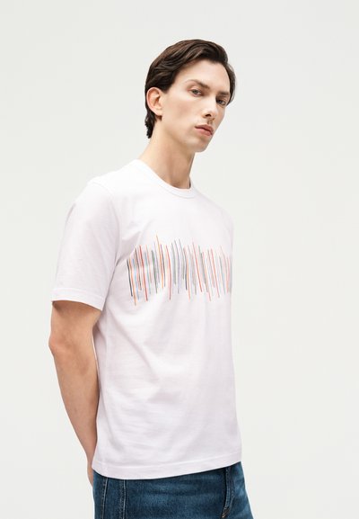 Νεαρός άνδρας που φοράει λευκό T-shirt με κάθετες πολύχρωμες γραμμές στο στήθος, συνδυασμένο με μπλε τζιν τζιν.