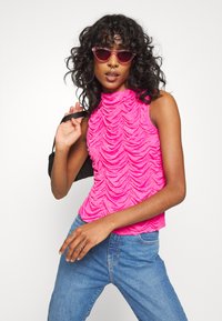 Top fúxia sem mangas com um decote alto, apresentando camadas de babados texturizados. Combinado com jeans azuis e uma mala preta de ombro.