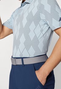 Polo bleu ciel avec un motif en losanges texturé, associé à un pantalon bleu marine et une ceinture grise. Coutures détaillées et patte de boutonnage visibles.