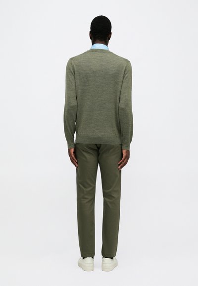 Uomo in piedi di spalle, vestito con un maglione verde oliva, pantaloni abbinati, colletto di camicia azzurro chiaro visibile e sneaker bianche.
