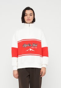 Λευκό φούτερ με κόκκινες λεπτομέρειες, ημιδιάπλακο κολάρο και κείμενο "Après Ski με εμένα". Διαθέτει γραφικό βουνού και ριγωτό σχέδιο.