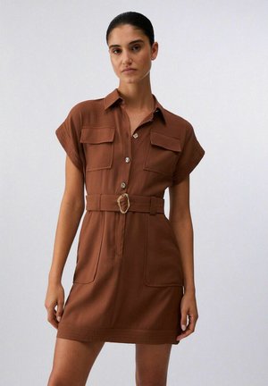 Donna che indossa un abito marrone a maniche corte con cintura, tasche frontali e dettagli con bottoni, in piedi davanti a uno sfondo chiaro uniforme.